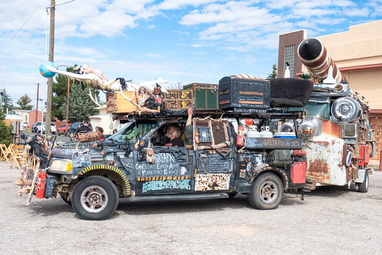 A decorated black pickup truck with chalk lettering, dolls and mannequins on the roof tows a rusty trailer topped with a striped cannon.