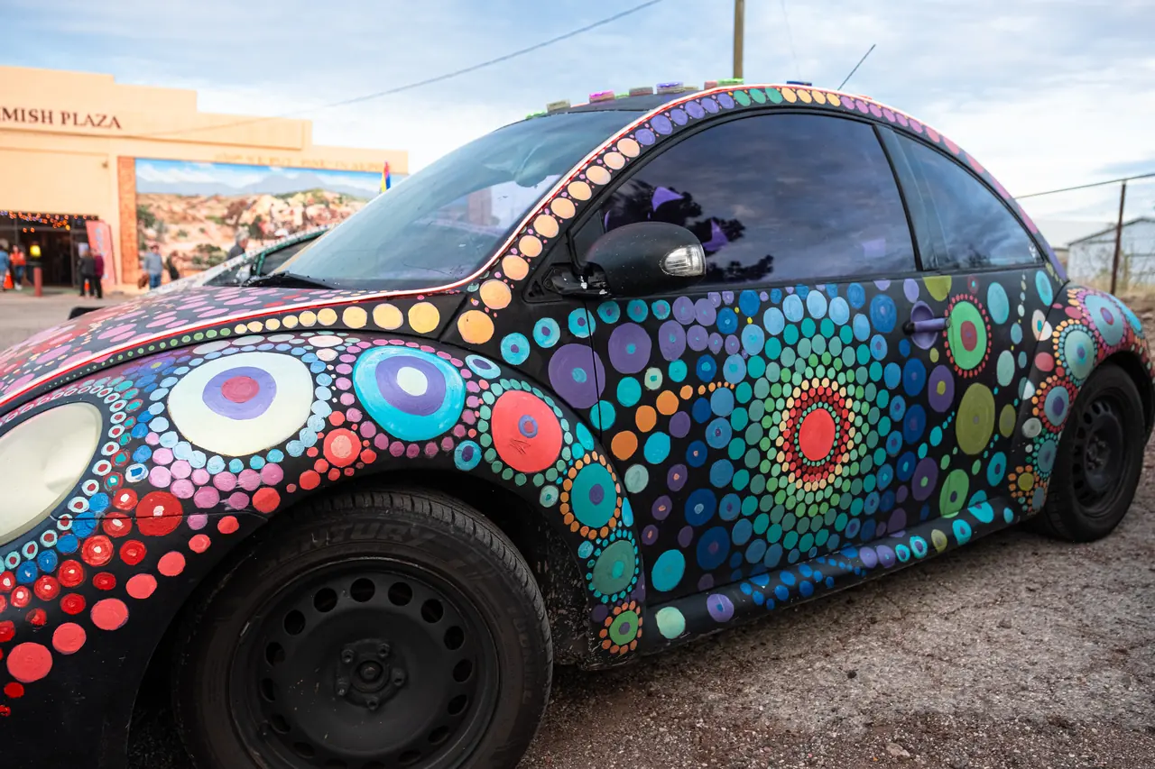 A black Volkswagen Beetle covered in colorful painted circles parked in front of a plaza building.