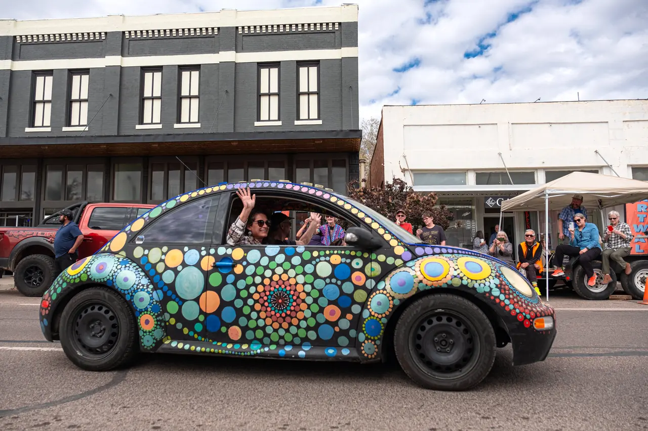 A black Volkswagen Beetle painted with colorful mandala dots drives down a street with passengers waving.