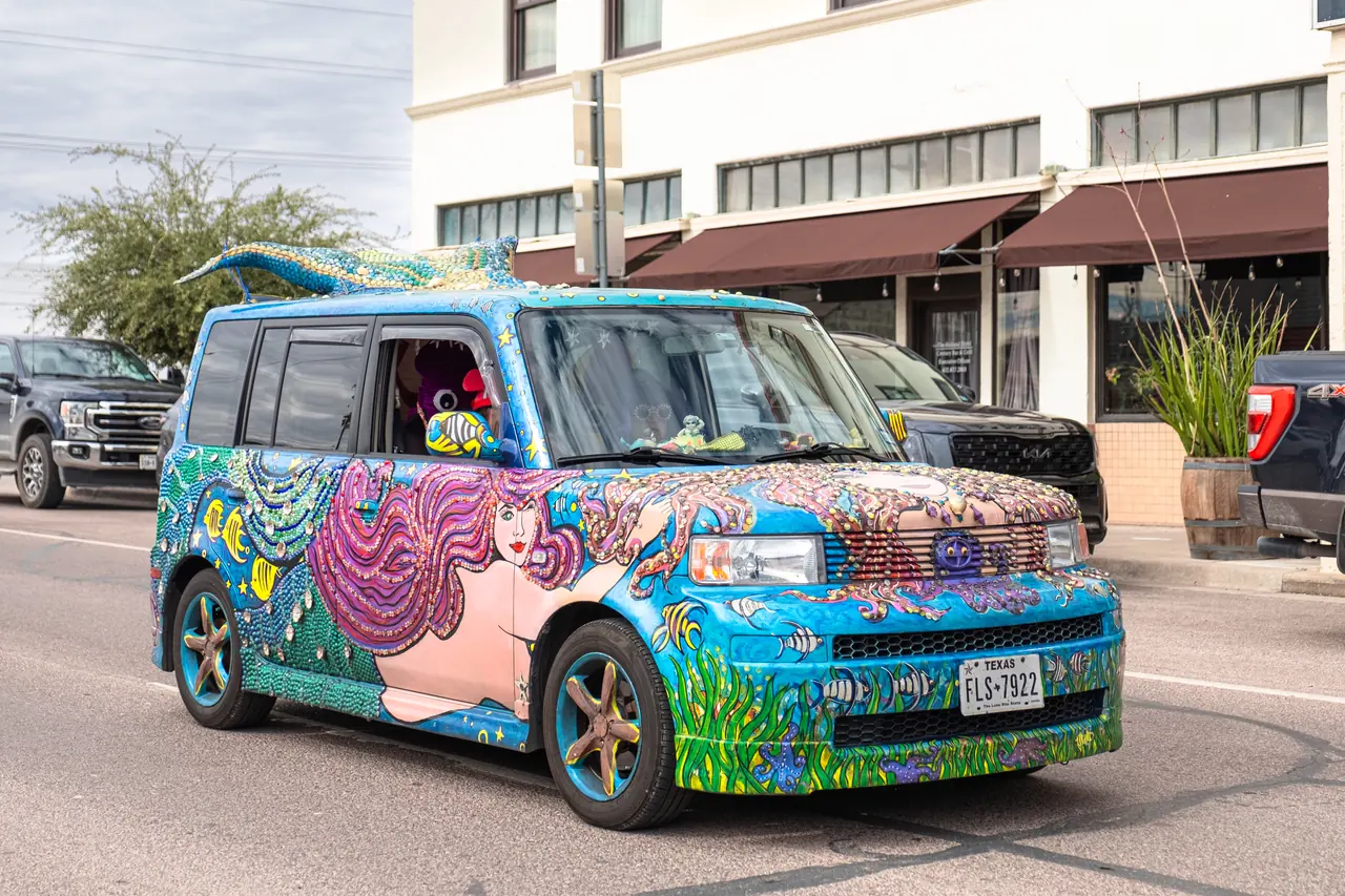 A blue box-shaped car covered in a painted mermaid, fish and beads with a sculpted mermaid tail on its roof.