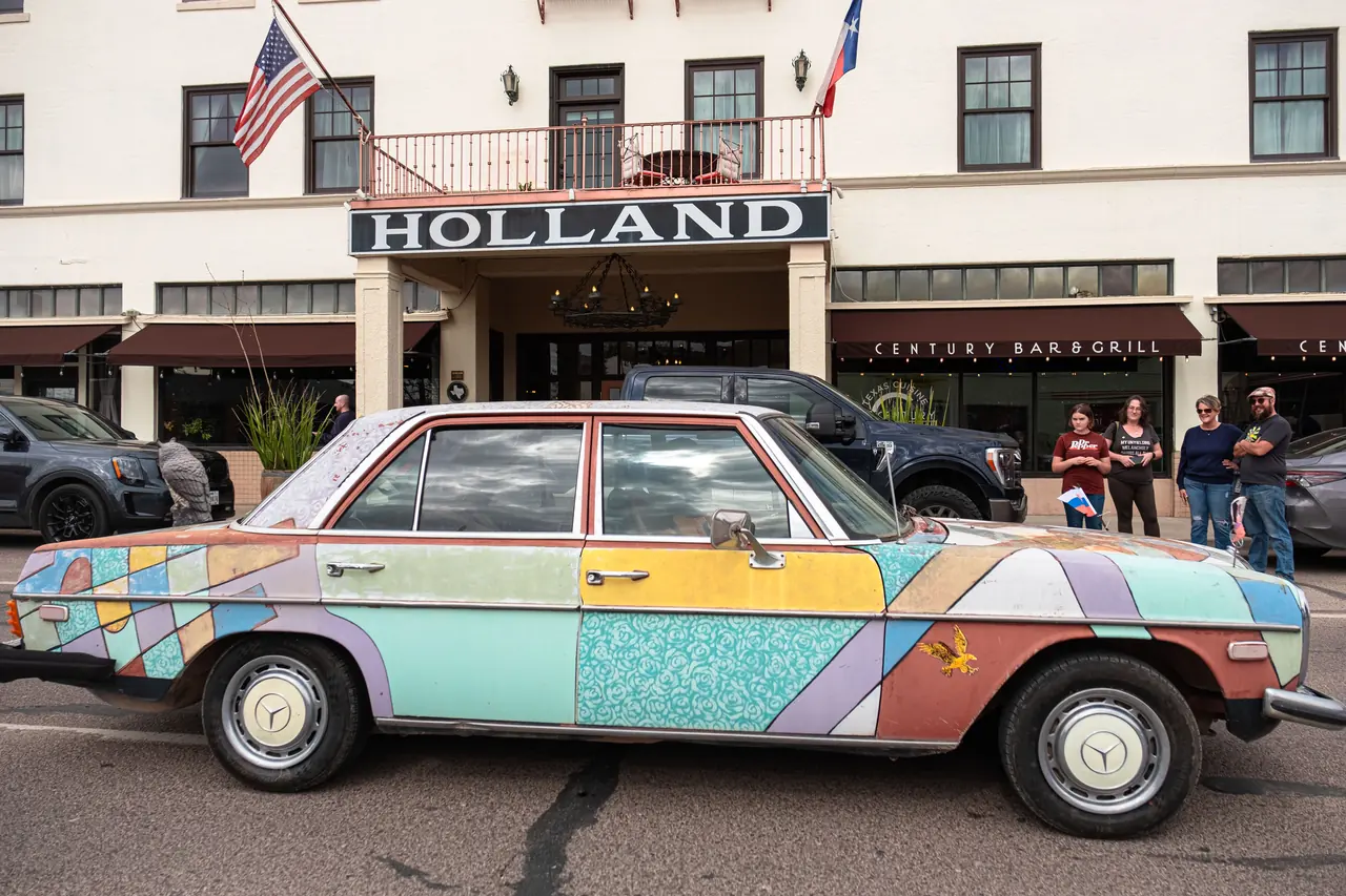 A vintage Mercedes sedan painted in pastel patchwork shapes parked on the street in front of a historic hotel.
