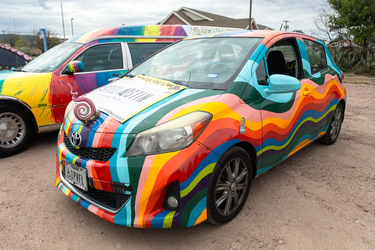 A small hatchback painted with flowing rainbow stripes parked on gravel beside another colorfully painted car.