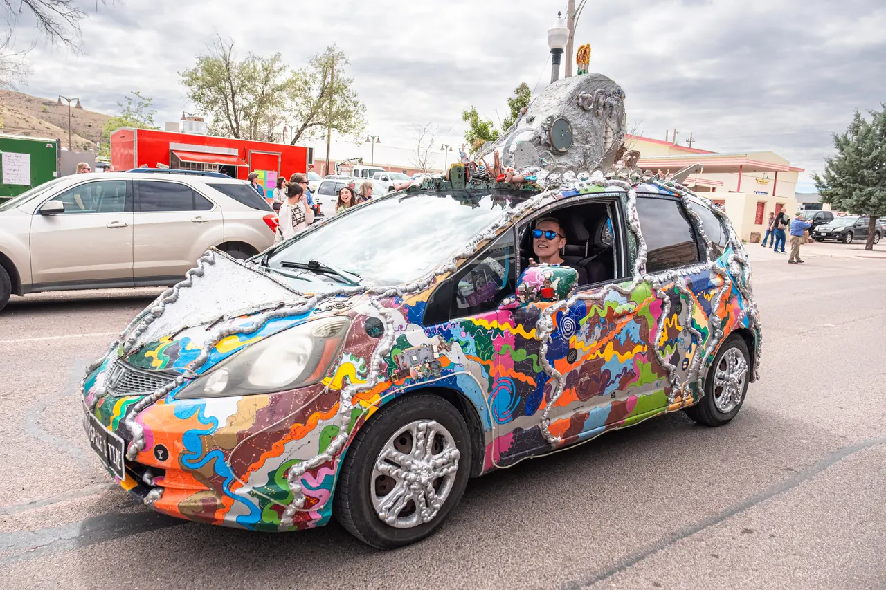 A small car covered in colorful painted patterns and silver sculpted tubing with a metallic creature sculpture on its roof.