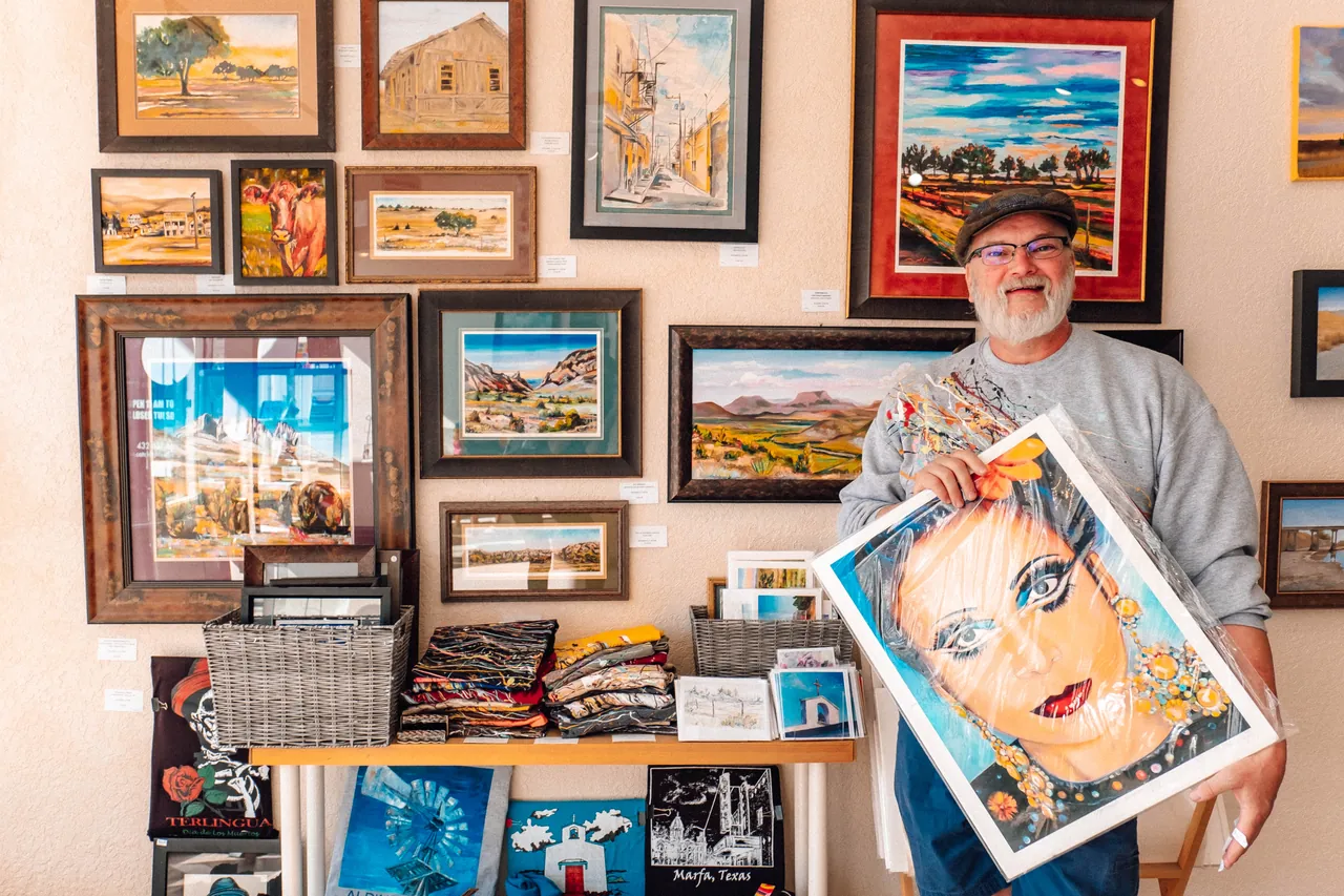An artist in a cap smiles in his gallery booth, holding a colorful portrait painting, with framed landscapes on the wall behind him.