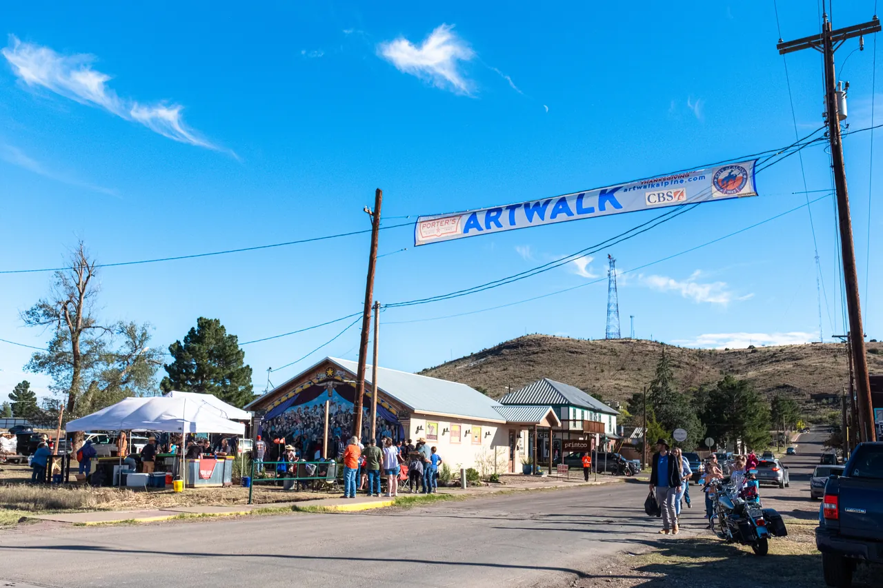 An Artwalk banner hangs across a street in Alpine under a blue sky, with festival tents, people and a hill behind.