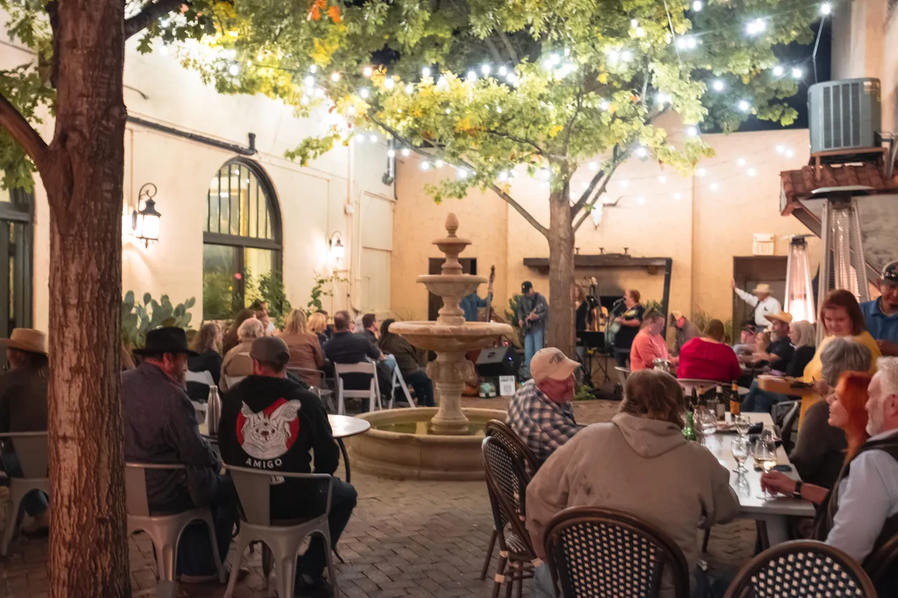 A courtyard patio with a fountain, filled with people at tables under string lights in the evening.
