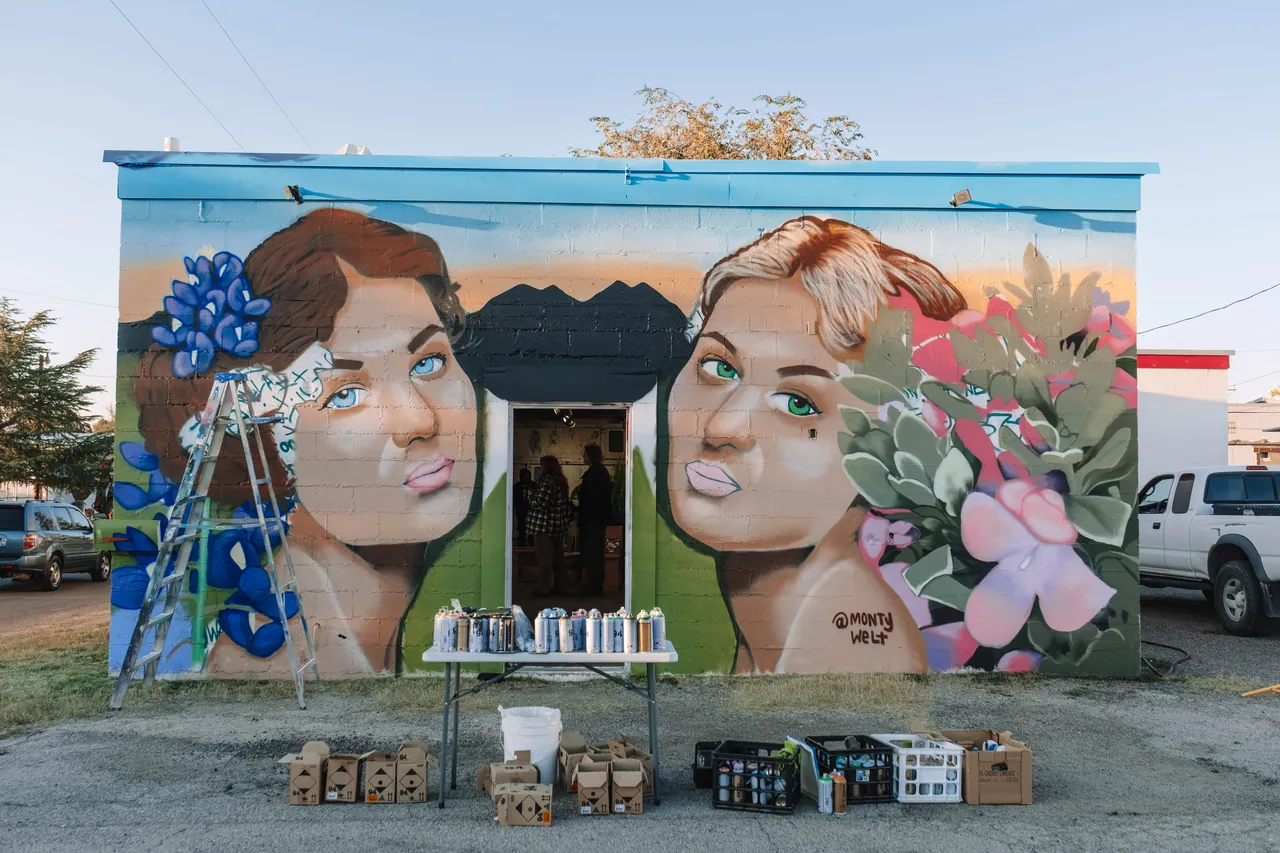 A colorful mural of two painted faces and flowers covers the side of a building, with paint cans on a table in front.