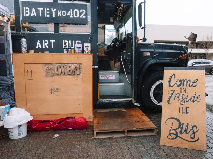 The Batey No. 402 Art Bus, a converted dark green school bus, with its door open beside a Come on Inside the Bus sign.